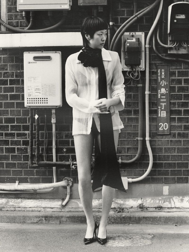 Monochrome portrait of a slender Asian woman in a semi-sheer white shirt, black scarf, and black heels posing against a brick wall in an urban setting.