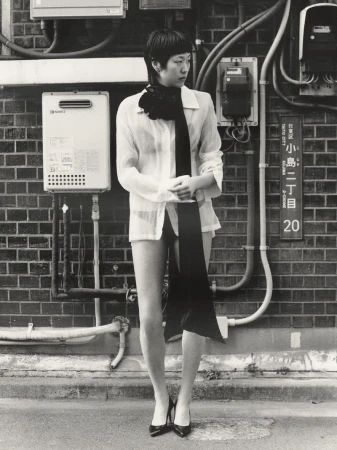Monochrome portrait of a slender Asian woman in a semi-sheer white shirt, black scarf, and black heels posing against a brick wall in an urban setting.