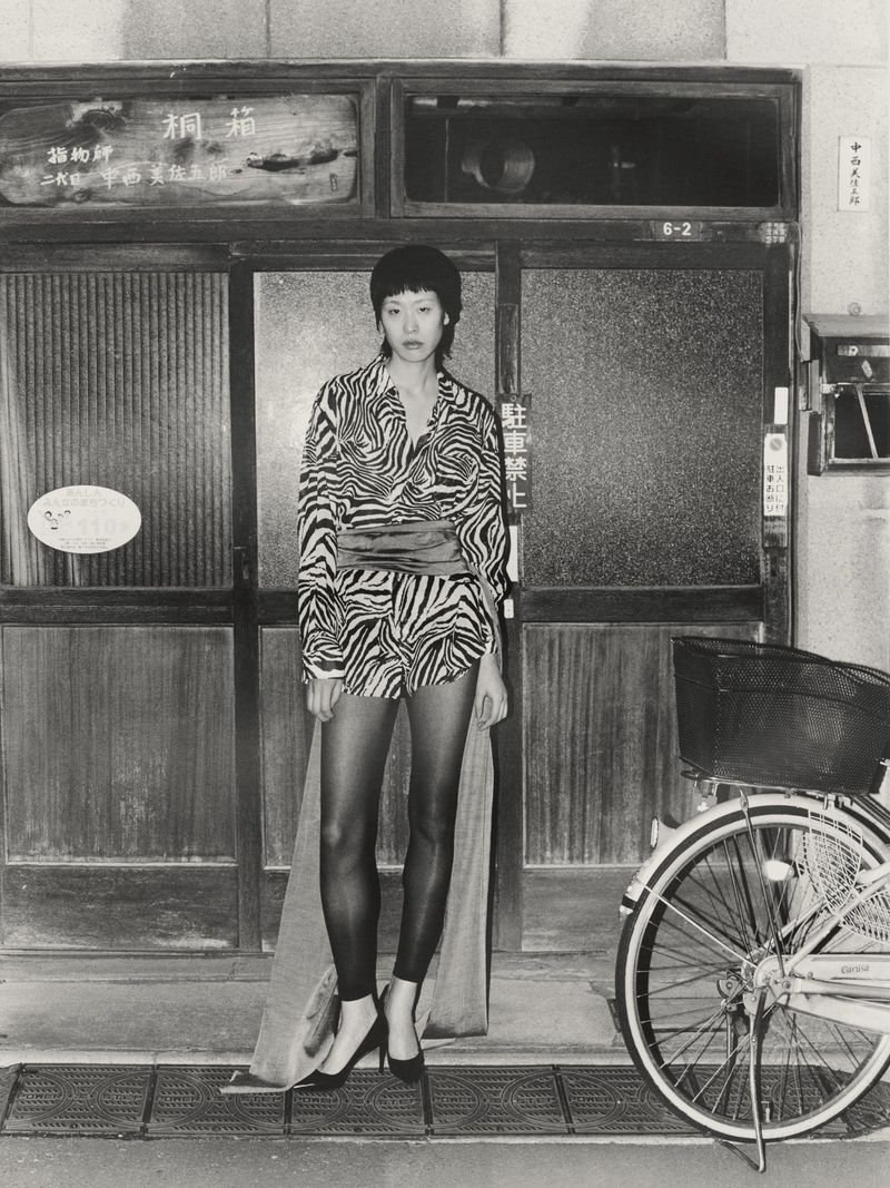 Monochrome portrait of an Asian woman in a patterned top and black tights, standing near a bicycle in front of a weathered doorway.