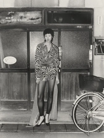 Monochrome portrait of an Asian woman in a patterned top and black tights, standing near a bicycle in front of a weathered doorway.