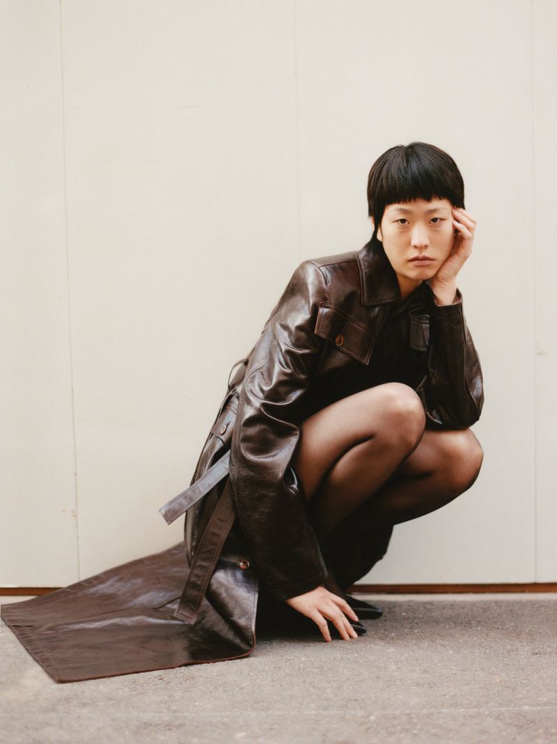 An adult female model poses in a dark leather trench coat against a light wall, giving a serious facial expression while crouching.