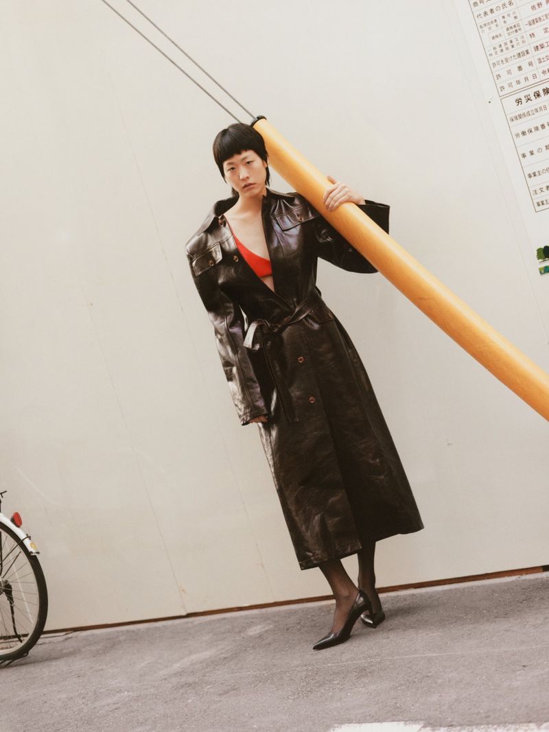An Asian woman in a black trench coat carries a large yellow pipe in an outdoor urban setting with a bicycle in the background.