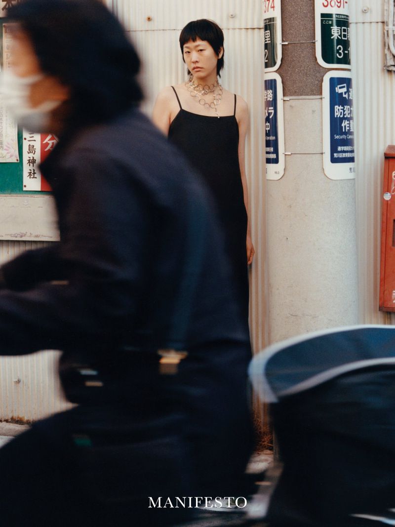 Woman in a black dress with a necklace stands on a city sidewalk near utility poles and signage; person blurred in foreground.