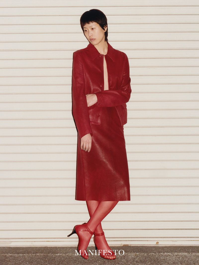 A woman in a red skirt suit and red tights poses against a corrugated metal wall in an eye-level medium shot, wearing matching heels.