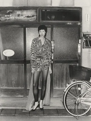 Model wearing patterned zebra print outfit standing in front of a traditional storefront with a bicycle parked alongside.