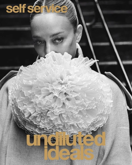 Black and white portrait of a woman with hair neatly styled back, wearing an oversized ruffled collar and standing near metal steps.