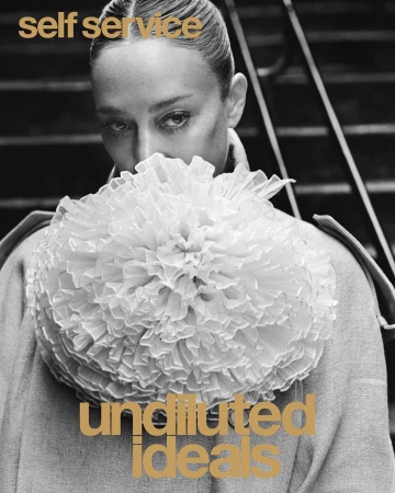 Black and white portrait of a woman with hair neatly styled back, wearing an oversized ruffled collar and standing near metal steps.