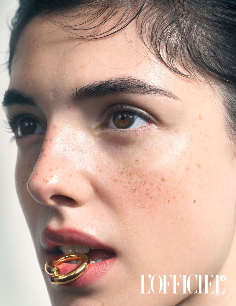 Close-up portrait of a woman with dark hair, freckles, and brown eyes holding a gold ring in her mouth against a white background.