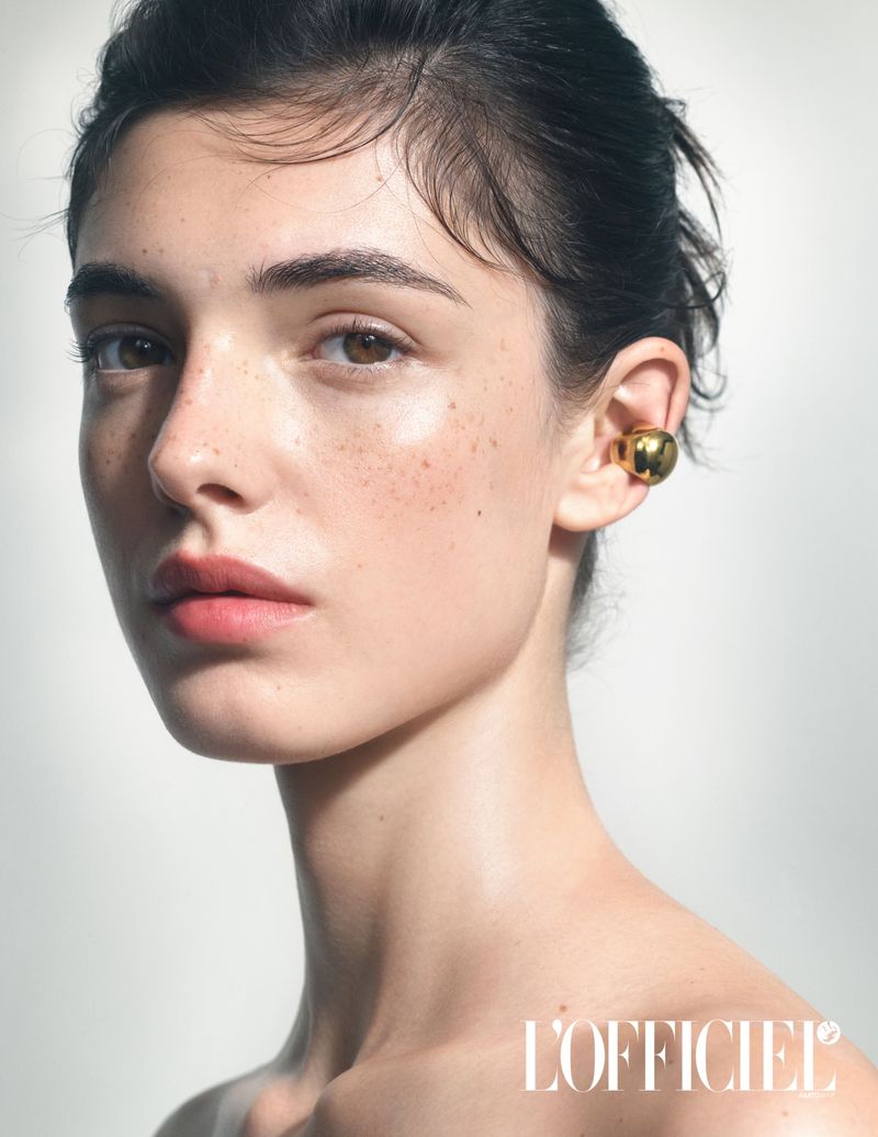 Close-up shot of a young woman with a gold ear cuff earring, fair skin, and dark hair, against a light background. Beauty fashion editorial.