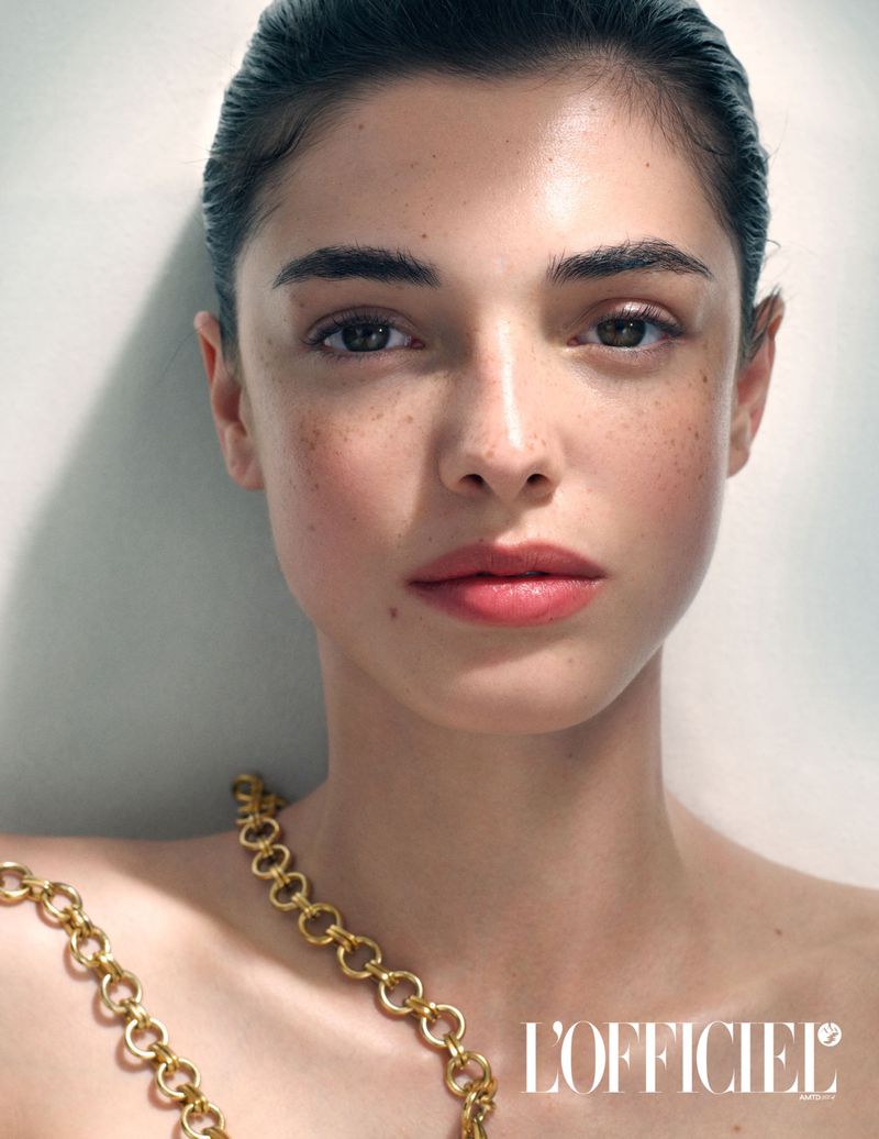 Close-up portrait of a beautiful woman with golden chain jewelry, light makeup, and freckles, posing in front of a light background.