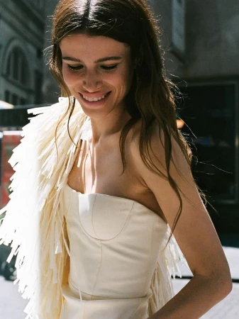Smiling woman in a strapless top and a fringed shawl outdoors, with a sunny backlight and blurred city buildings in the background.