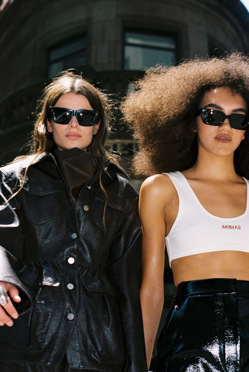 Two women pose in an urban setting wearing sunglasses, leather jackets, and fashionable tops, showcasing contemporary street style.