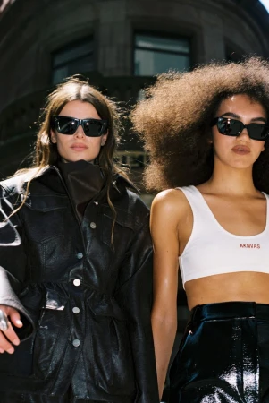 Two women pose in an urban setting wearing sunglasses, leather jackets, and fashionable tops, showcasing contemporary street style.