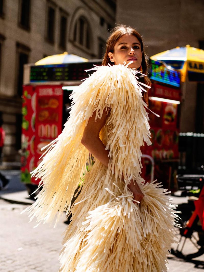 Woman poses in an avant-garde dress made of straw in an urban setting with buildings and food carts in the background.