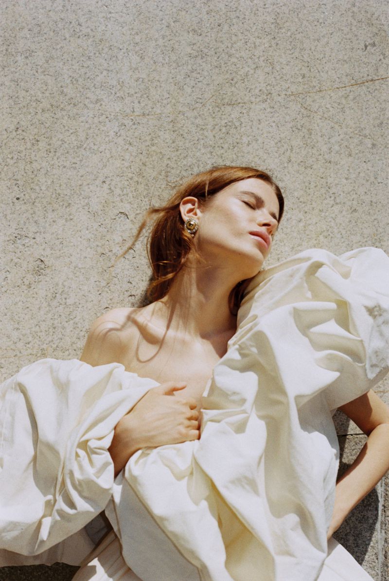 Woman with closed eyes wearing a voluminous off-white ruffled dress resting against a textured stone surface in bright sunlight.