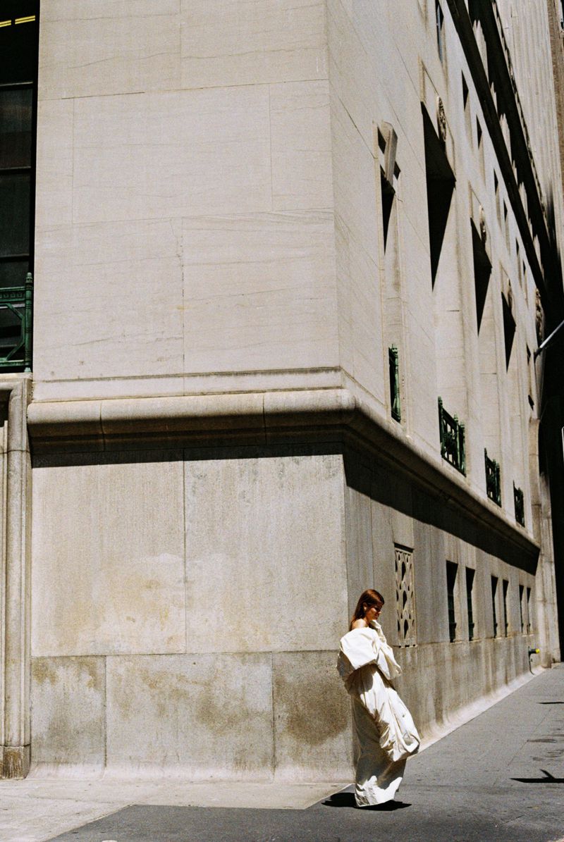 Woman in a white dress walks along a city street next to a tall stone building with geometric windows and bright sunlight, fashion photography.