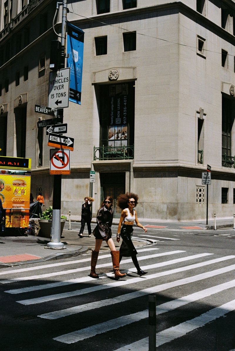 Two women in fashionable attire cross a zebra-striped crosswalk in an urban setting with vintage styling. Pedestrians, buildings and signs visible.