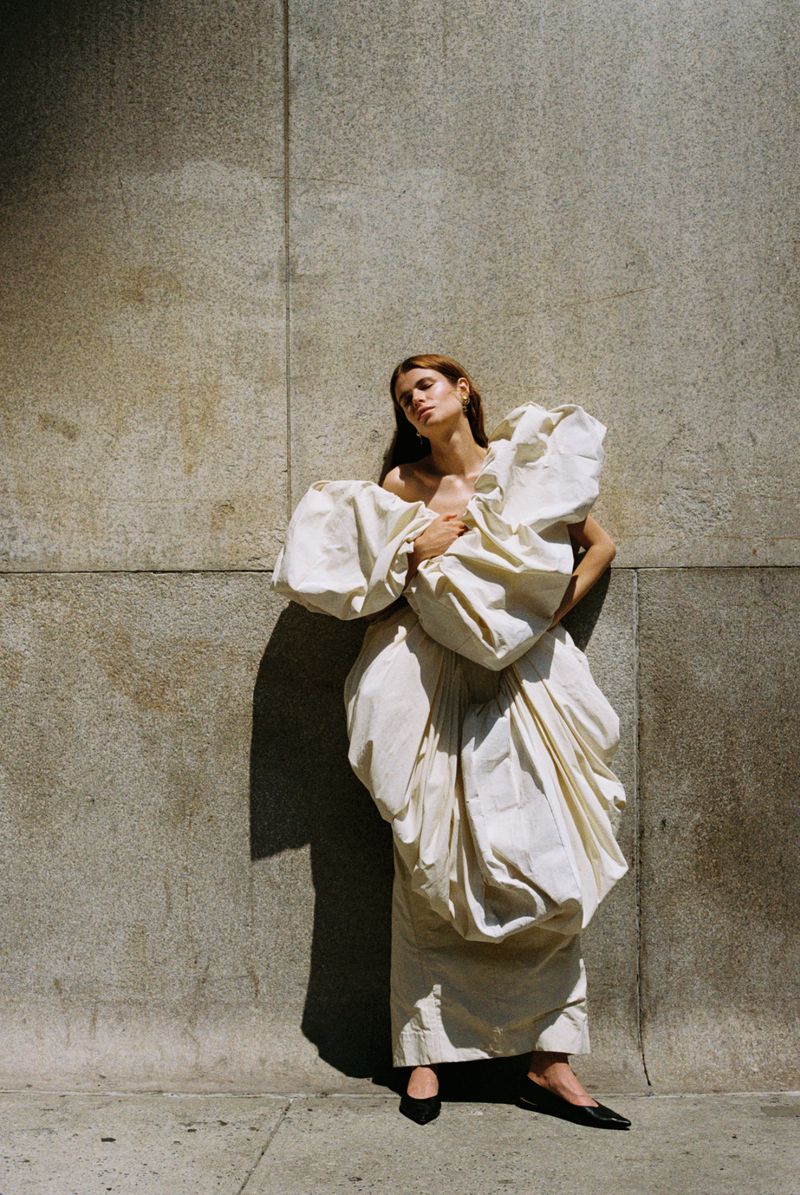 Woman in oversized draped dress poses against concrete wall in natural light. Fashion shot showcasing dramatic design and urban setting.