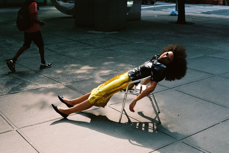 Woman with afro rests on metal chair outdoors on concrete. She wears a black jacket and gold skirt, with a man walking in the background.