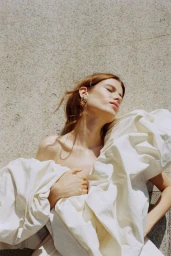 Woman with closed eyes wearing a voluminous off-white ruffled dress resting against a textured stone surface in bright sunlight.