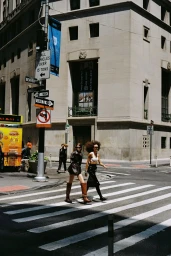 Two women walking across a city street crosswalk near a classic stone building and street signs for Tidal Magazine project Around Town.