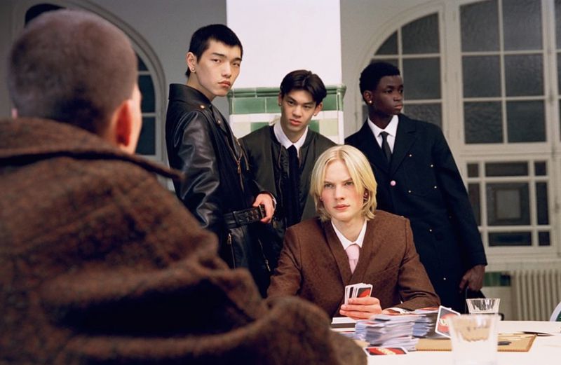 A group of five young adult men gather around a table, one holding playing cards in an indoor setting with large windows.