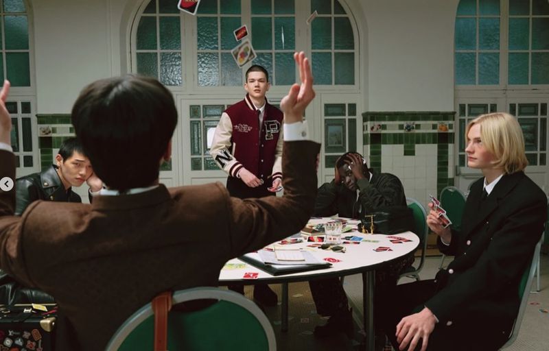 A group of five young adults gather around a table in a vintage-style room, playing cards, with cards flying in the air.