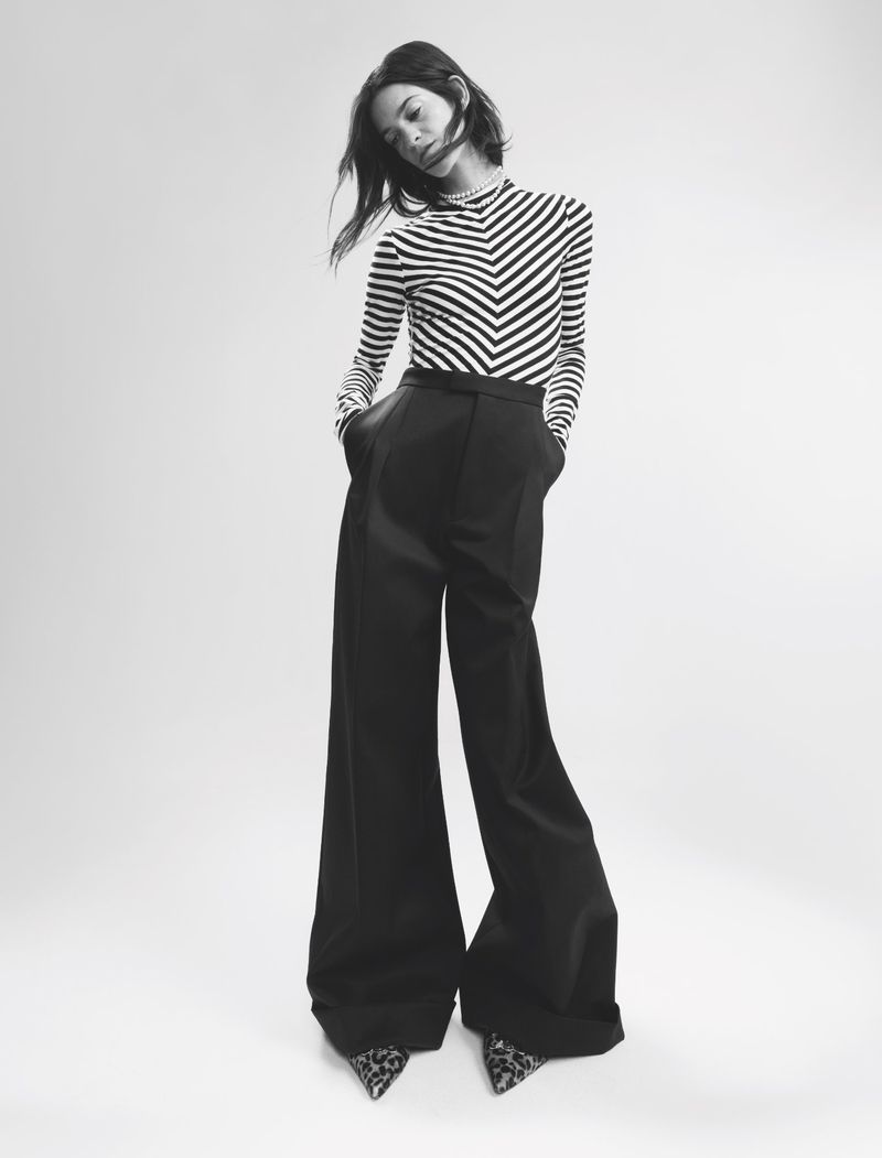 A young woman poses in a black and white chevron-striped top and black, wide-leg trousers in a studio environment with soft lighting.