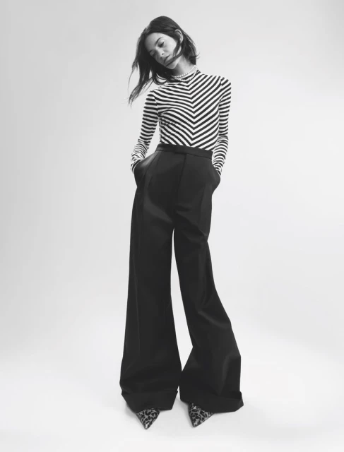 A young woman poses in a black and white chevron-striped top and black, wide-leg trousers in a studio environment with soft lighting.