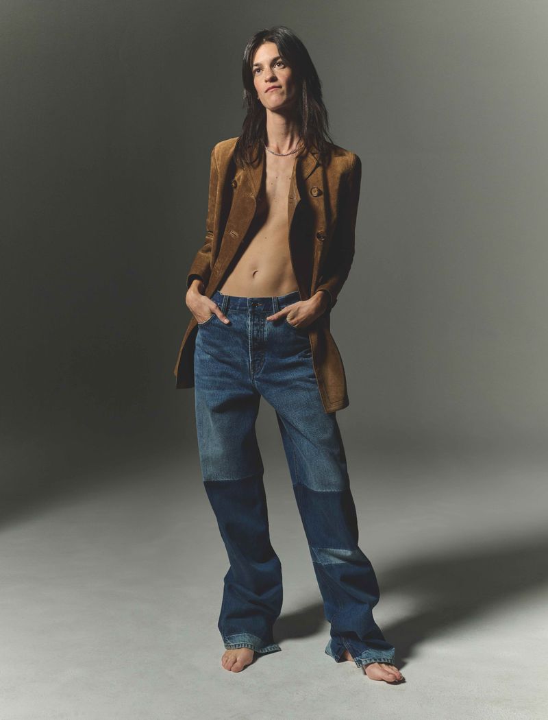 A woman in jeans and a brown blazer stands barefoot in a studio setting, looking confidently at the camera with neutral facial expression.