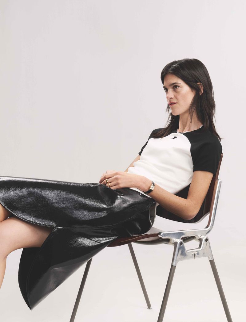 A brunette woman sits on a metal chair in a studio, wearing a white and black short-sleeved shirt and a shiny, black, flared skirt.