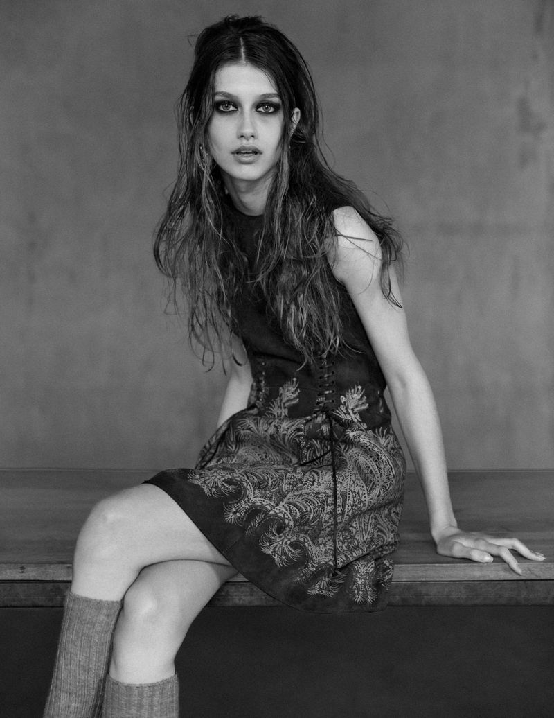 Monochrome portrait of a young woman with long dark hair, wearing a patterned dress and knee-high socks, sitting indoors.
