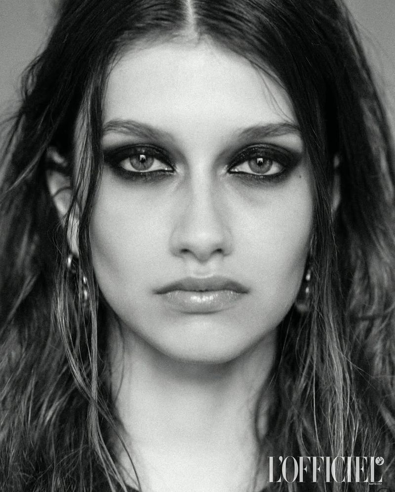 Close-up portrait of a young woman with dark hair and dramatic black eye makeup, silver hoop earrings, and neutral expression, black and white.
