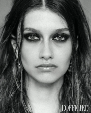 Close-up portrait of a young woman with dark hair and dramatic black eye makeup, silver hoop earrings, and neutral expression, black and white.