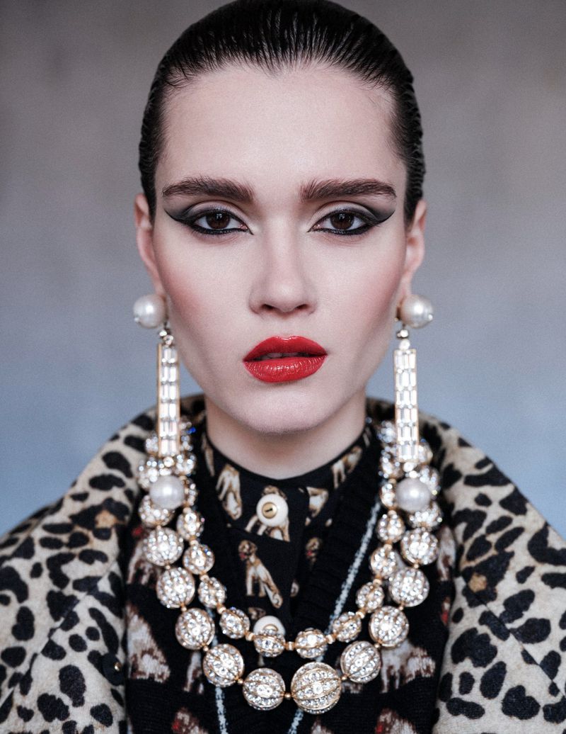 Fashion portrait of a woman with bold eye makeup, red lipstick, and large pearl earrings and necklace posing in front of a gray background.