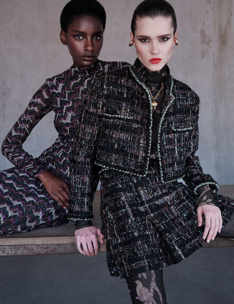 Two women pose wearing tweed jackets and skirt. One woman has an African complexion.  High fashion shot, studio background, close up.