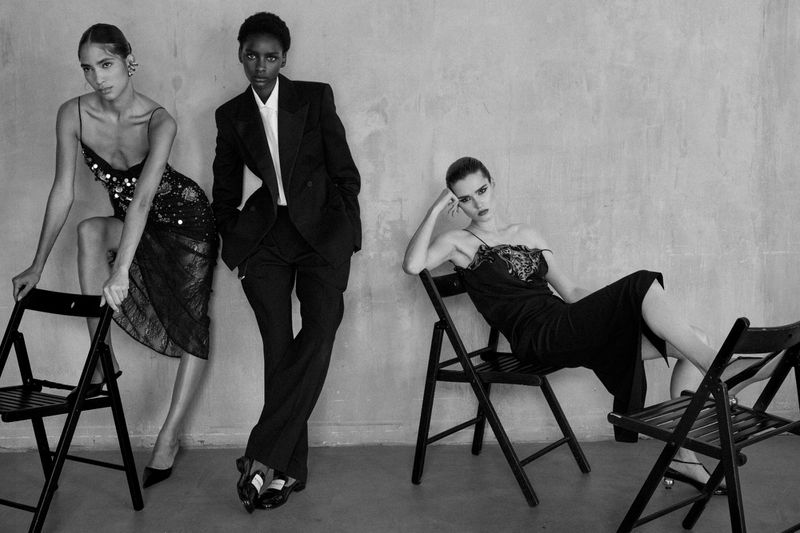 Three fashion models pose in black dresses and a suit, leaning on chairs against a neutral backdrop in a high-contrast black-and-white photograph.