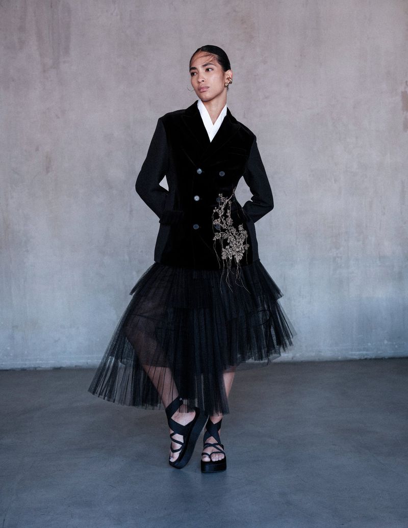 Elegant woman poses in a black velvet blazer, tulle skirt, and platform sandals against a neutral background in a studio fashion shot.