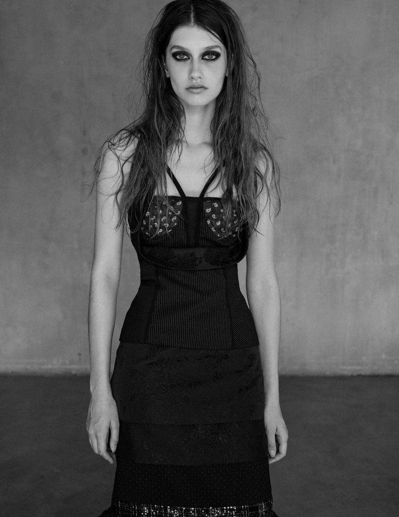 Black and white portrait of a woman with long, wavy hair, wearing a black dress, posing against a textured grey background in a studio setting.