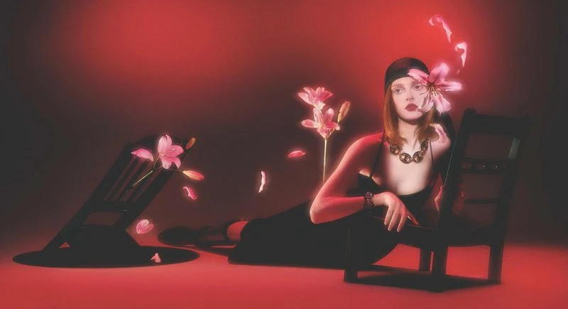 A woman in a black dress reclines on a chair in a studio setting with a red backdrop and pink flowers.
