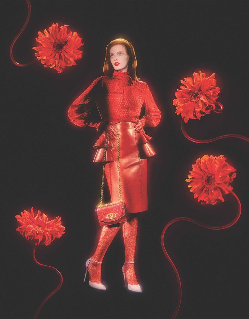 Fashion model wearing a red skirt and blouse, posing with a purse and dahlia flowers against a dark background in a studio shot.