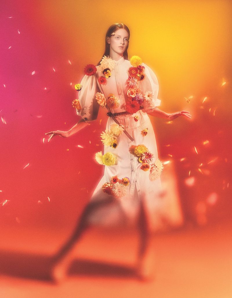 Woman in white dress with floral embellishments poses against a vibrant orange and pink background, creating a surreal, dreamlike fashion image.