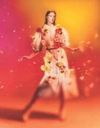 Woman in white dress with floral embellishments poses against a vibrant orange and pink background, creating a surreal, dreamlike fashion image.