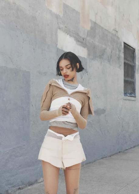 Young woman in casual fashion poses against a textured concrete wall, wearing shorts, top, and bolero in neutral tones, street style.
