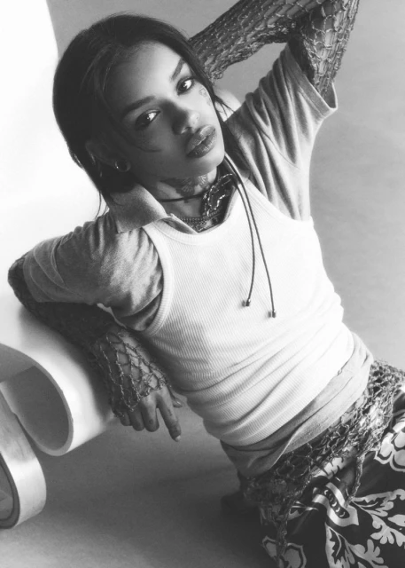 Portrait of an adult woman wearing a tank top and fishnet sleeves, sitting indoors, resting on a white sculptural object in black and white.