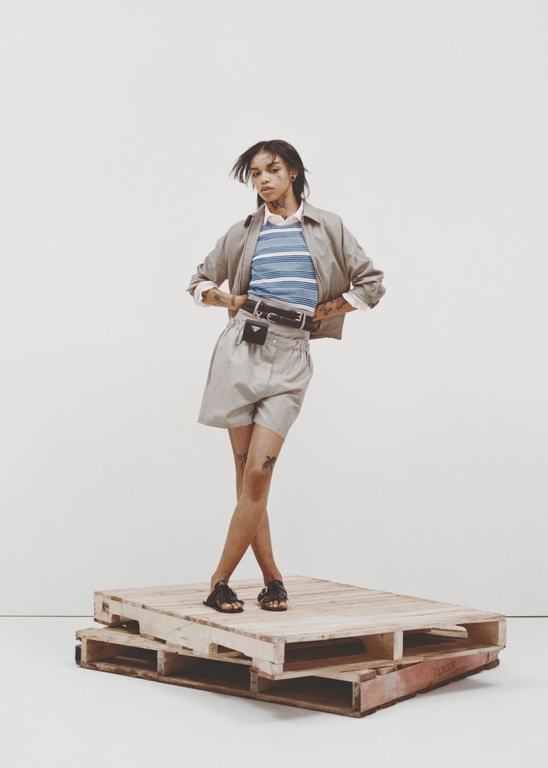 An adult female poses in a beige outfit, including a jacket, shorts, and sandals, on a wooden platform against a light background.