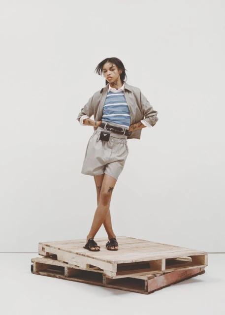 An adult female poses in a beige outfit, including a jacket, shorts, and sandals, on a wooden platform against a light background.