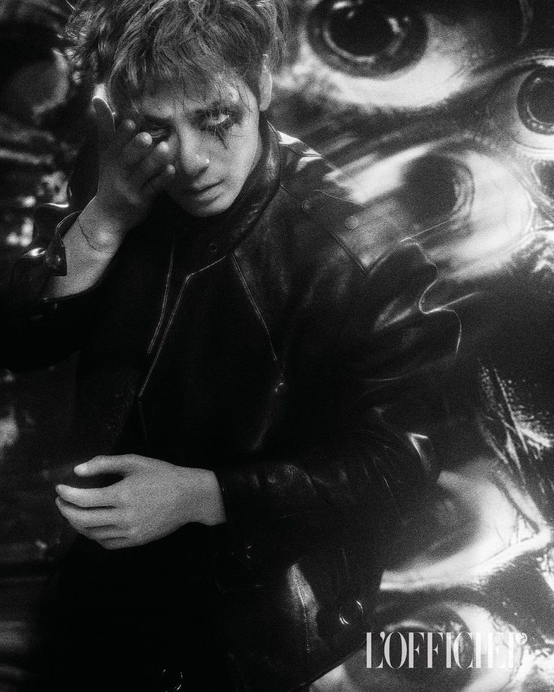 Monochrome portrait of a young man with theatrical makeup wearing a black leather jacket, posing with a blurred, surreal eye background.
