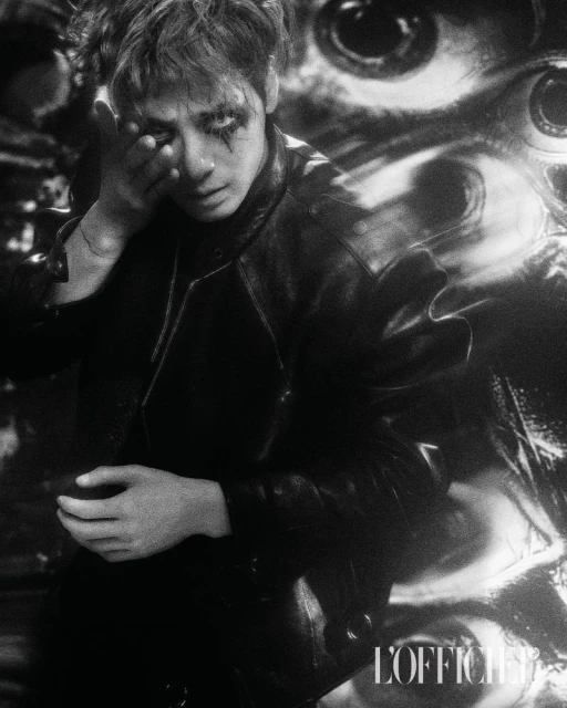 Monochrome portrait of a young man with theatrical makeup wearing a black leather jacket, posing with a blurred, surreal eye background.