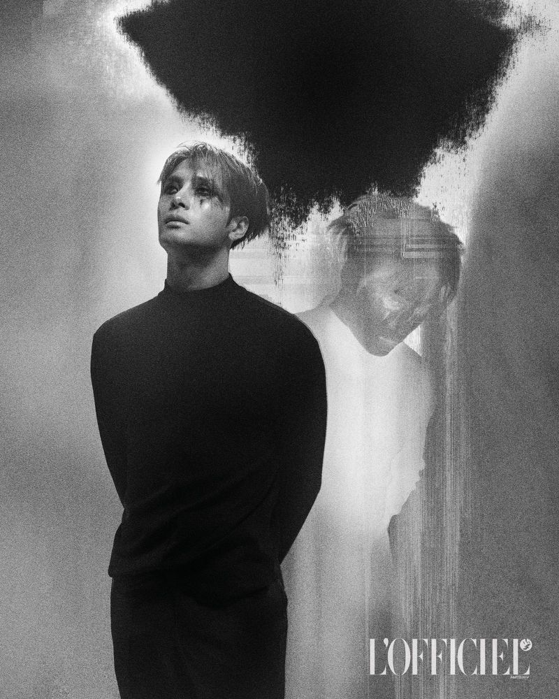 Monochrome studio portrait of an adult male with dark eye makeup looking up, accompanied by a blurry ghosted figure and black ink blot.